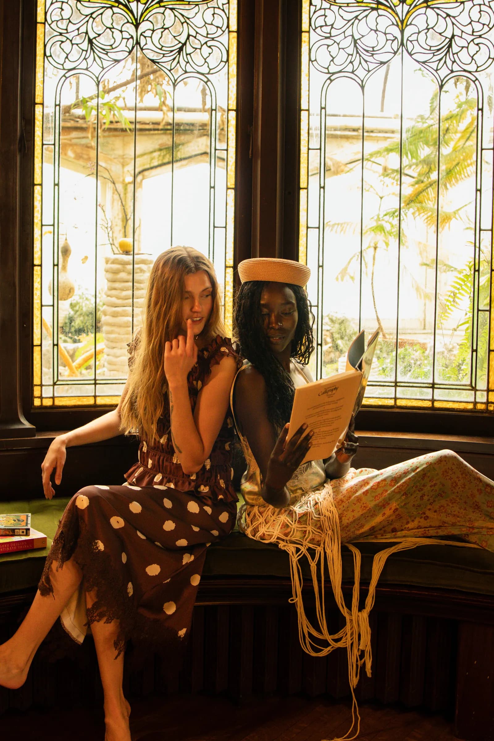 Two women sit close on a green bench in front of stained glass, one reading over the other's shoulder.