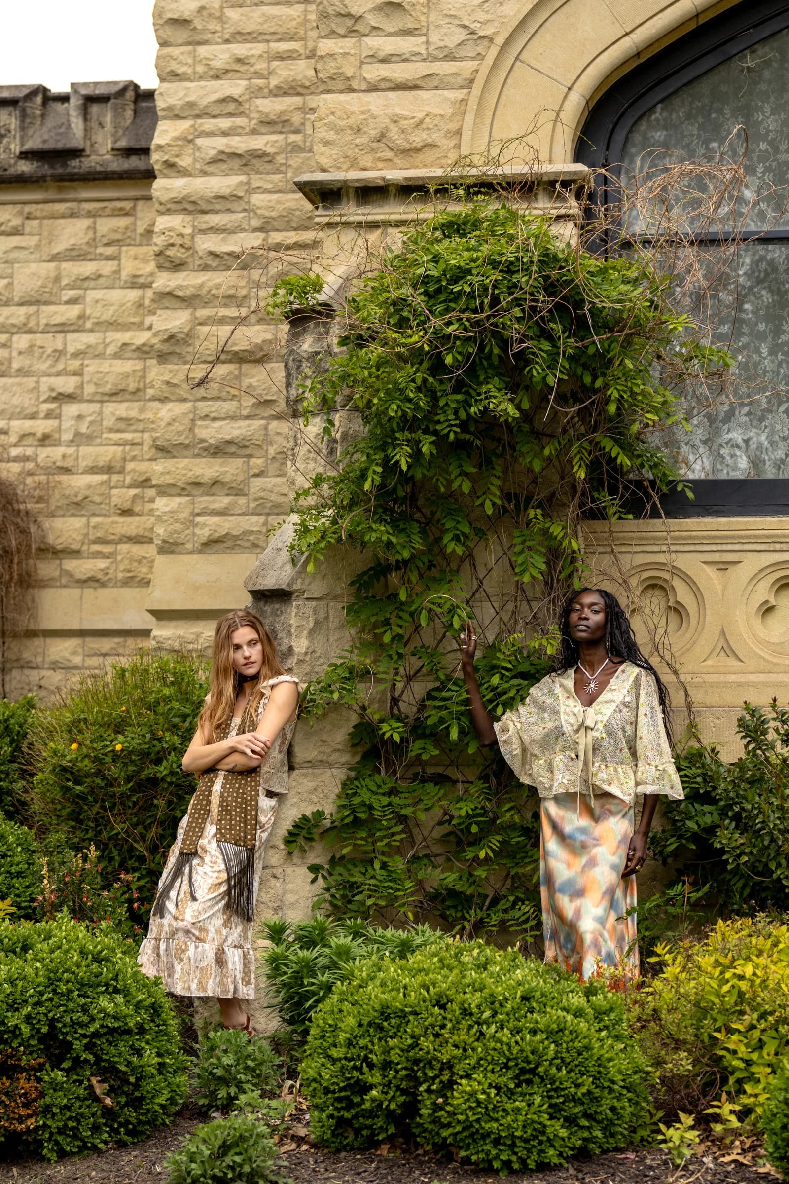 Two women stand against an ivy-covered stone wall beneath a tall arched window.