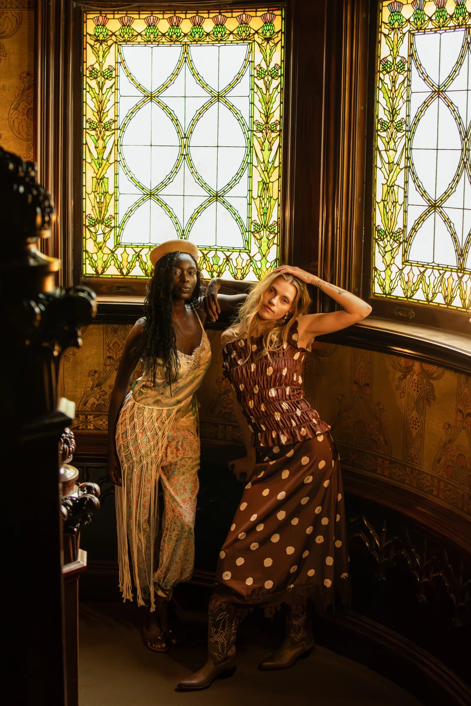 Two women lean into each other beneath two stained-glass windows at the top of a wooden staircase.