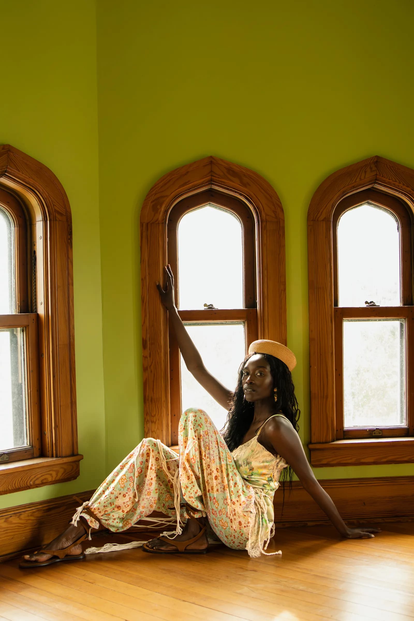 A woman sits on a sunlit wooden floor in a green room with three arched windows.