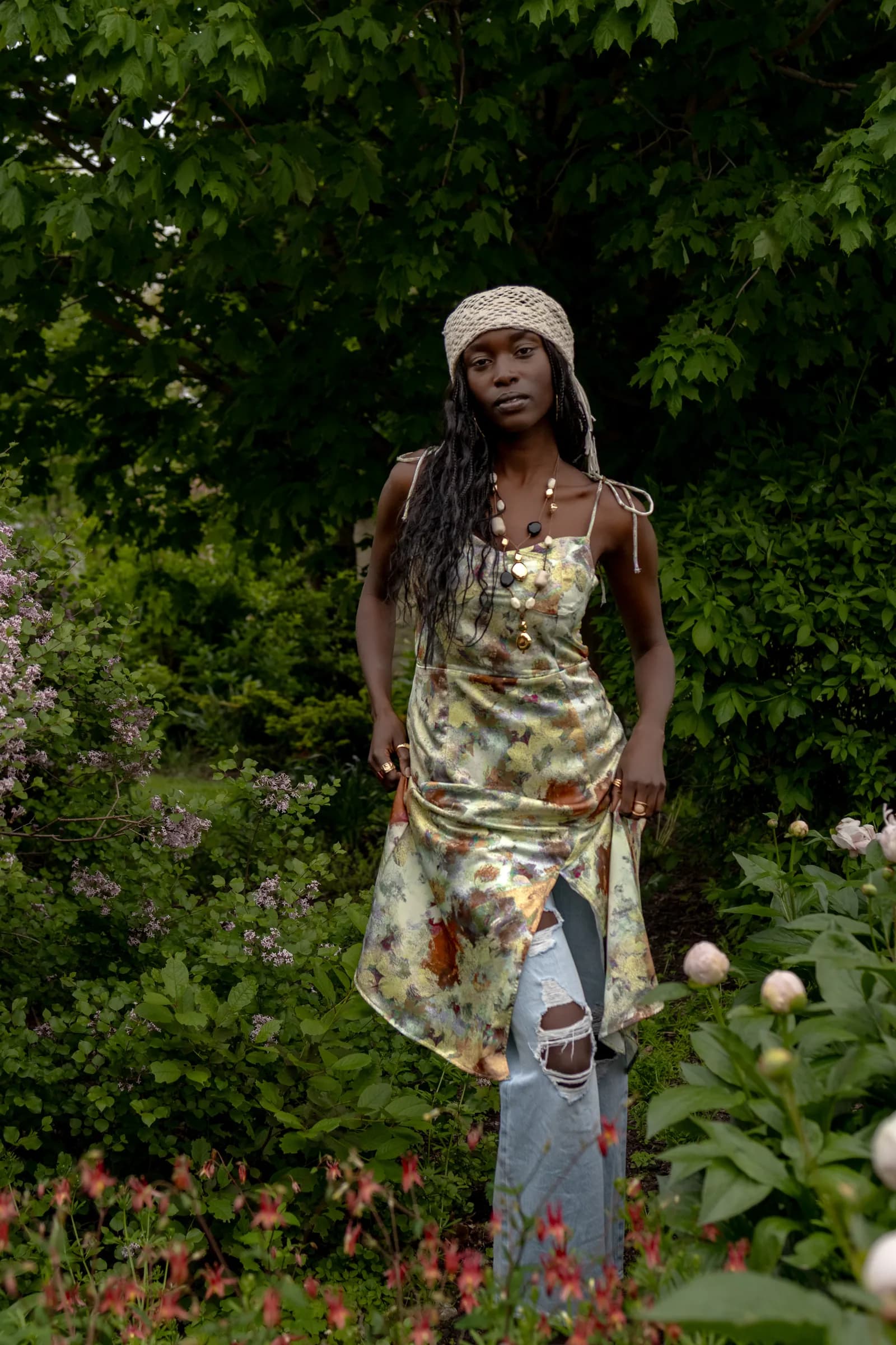 A woman in a headscarf and a floral slip dress stands among garden greenery.
