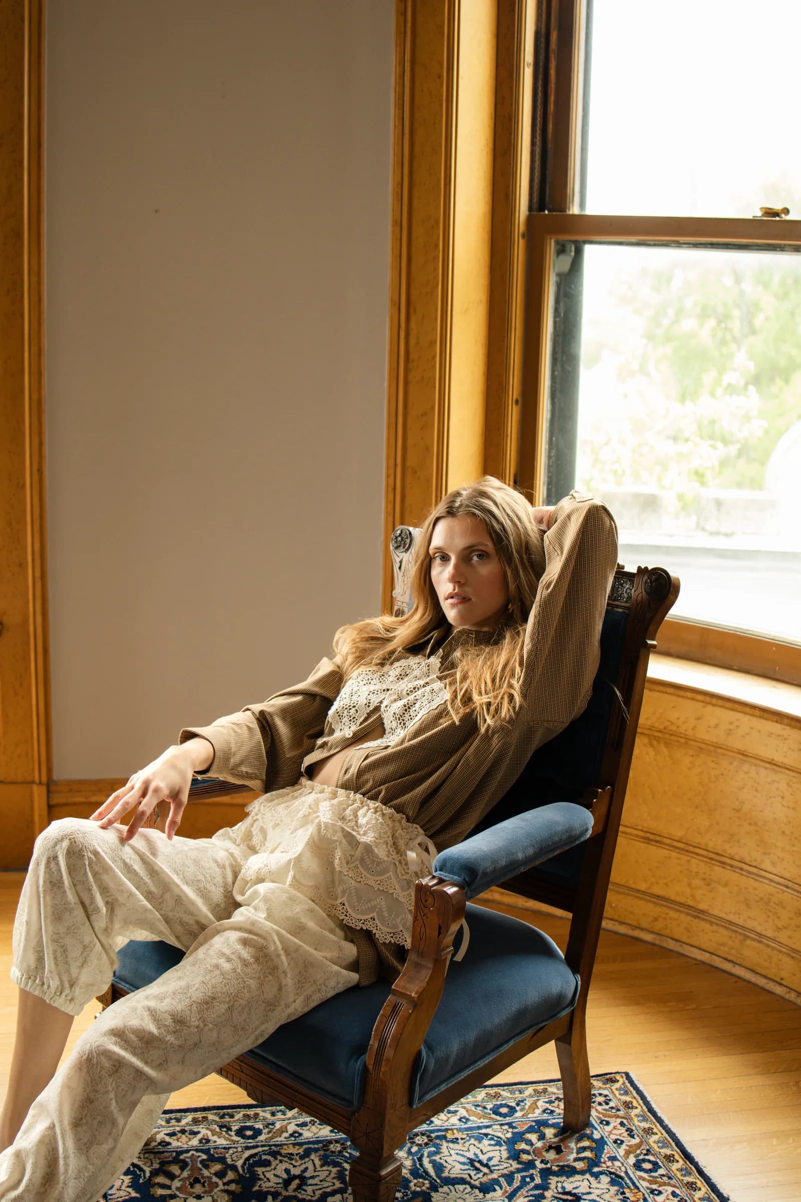 A woman in a tan shirt and lace-trimmed trousers sits back in a blue velvet chair by a window.