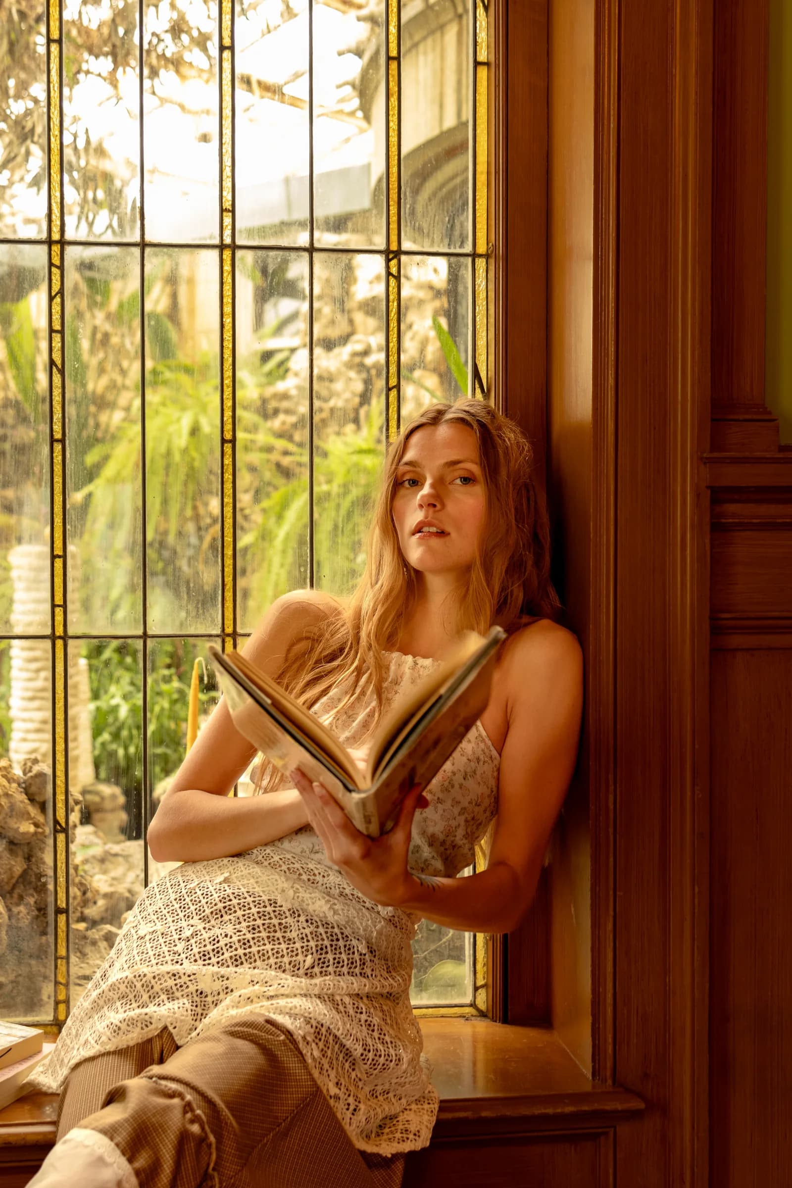 A woman sits in a window seat with a book, stained glass and greenery behind her.