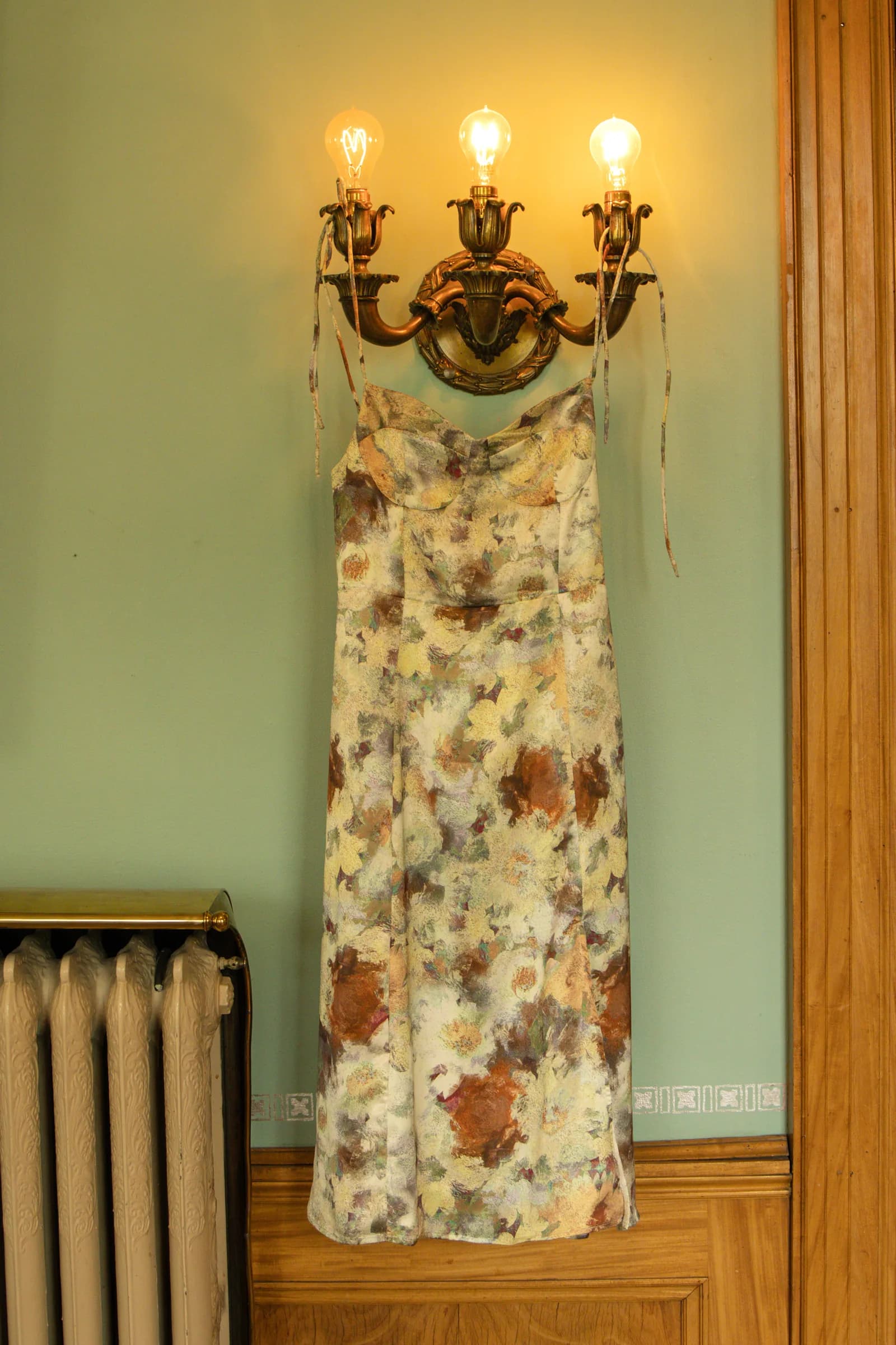 A floral dress hangs from a brass sconce on a pale green wall.