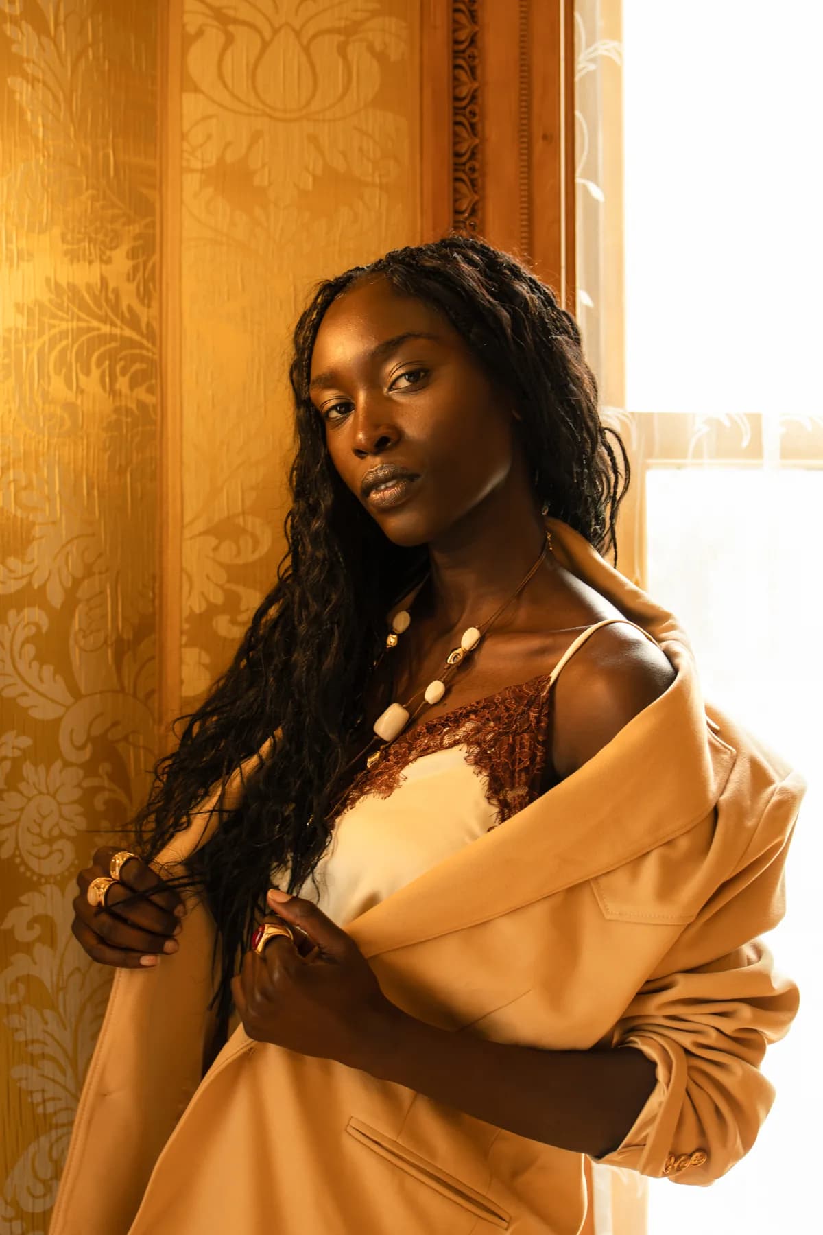 a woman by a sunlit window in an oversized camel blazer slipping off one shoulder over a cream satin tunic with brown lace