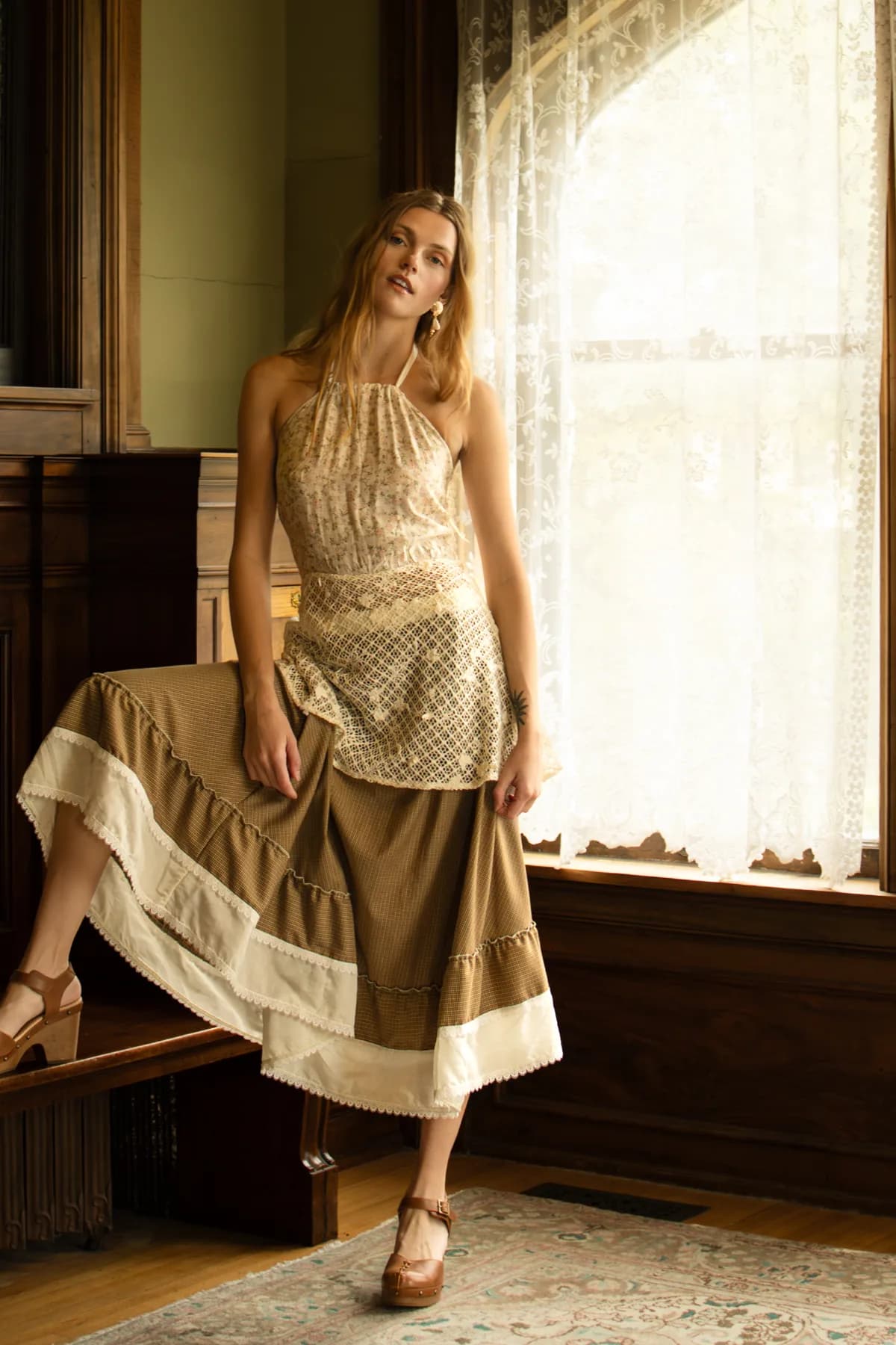 a woman by lace-curtained windows in a floral halter babydoll top and a brown check circle skirt with platform heels