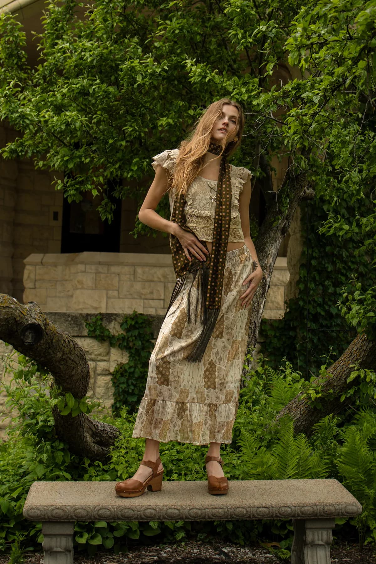 a woman standing on a stone bench in a garden in a patchwork top and matching midi skirt with a long skinny scarf and platform clogs