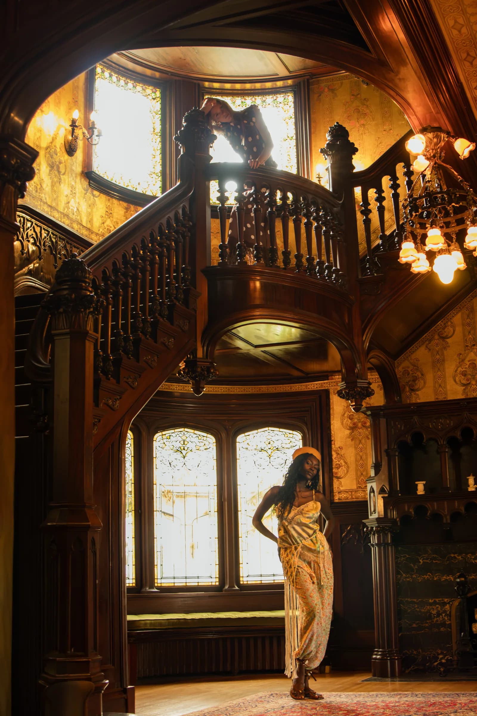 A woman stands at the foot of a grand wooden staircase lit by a stained-glass door.