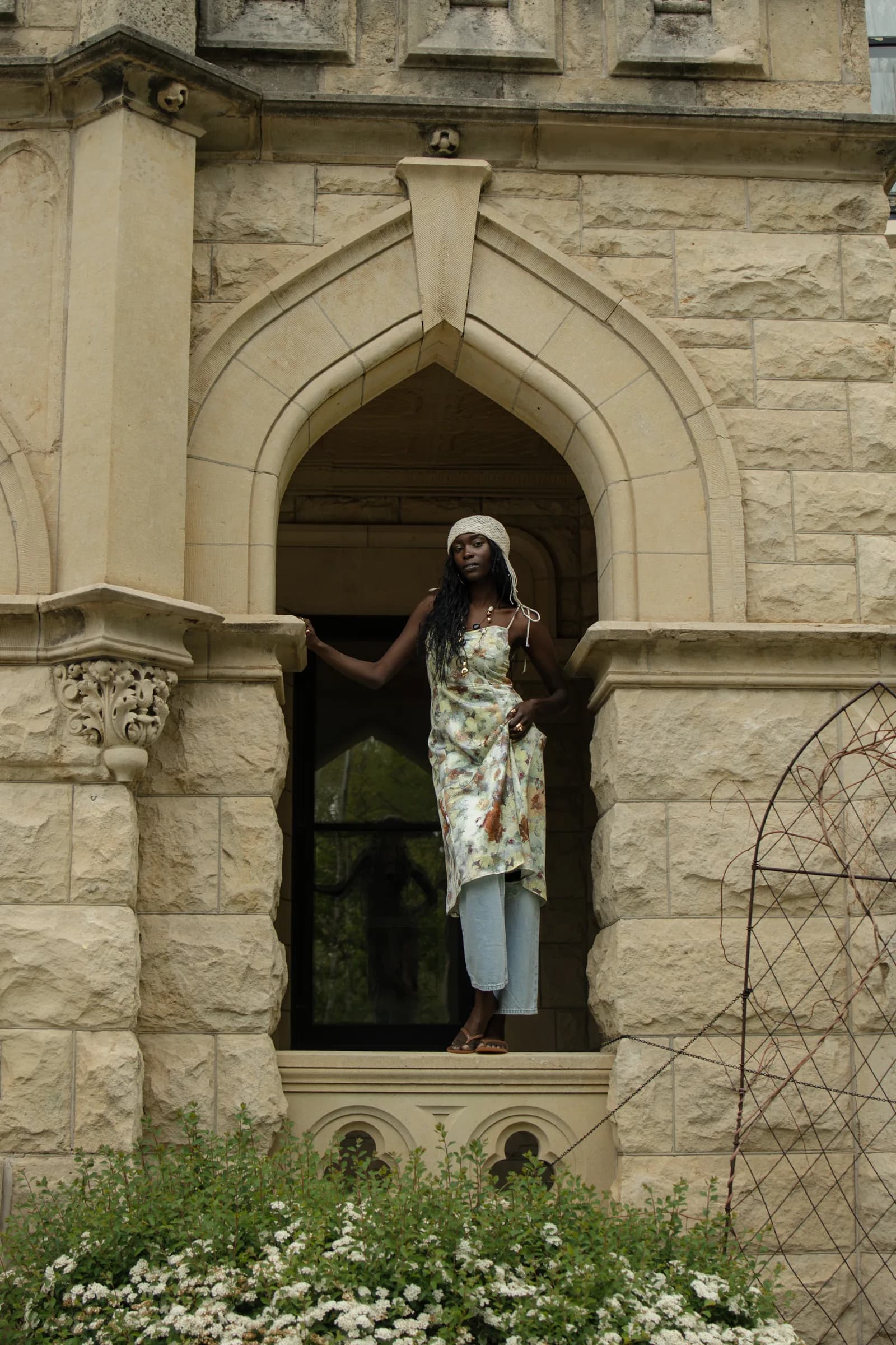 A woman stands framed in a carved stone arch above a bank of white flowers.