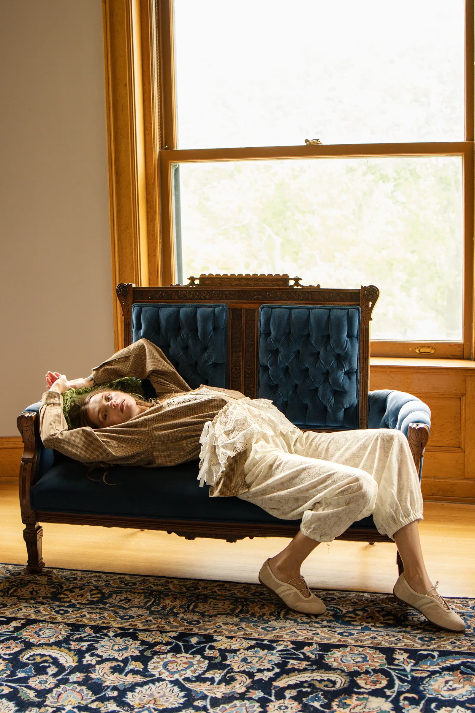 A woman stretches out across a blue velvet sofa in a bright parlour.