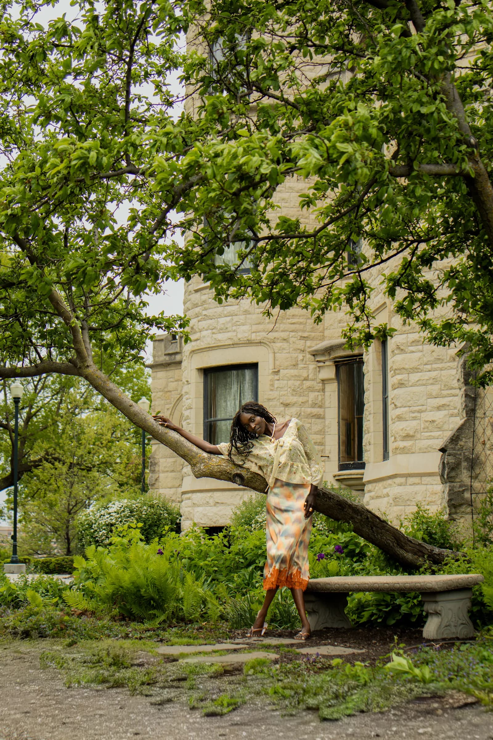 A woman perches on a low branch in the garden below the stone walls of the castle.