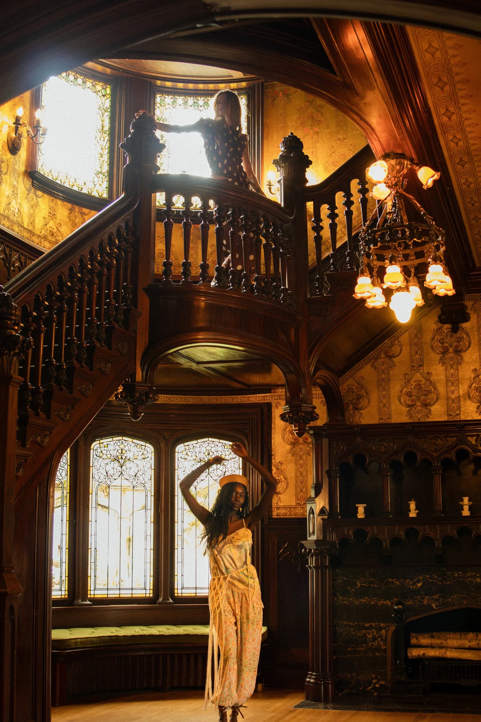 A woman stands at the foot of a carved wooden staircase, lit by a chandelier and a leaded glass door.