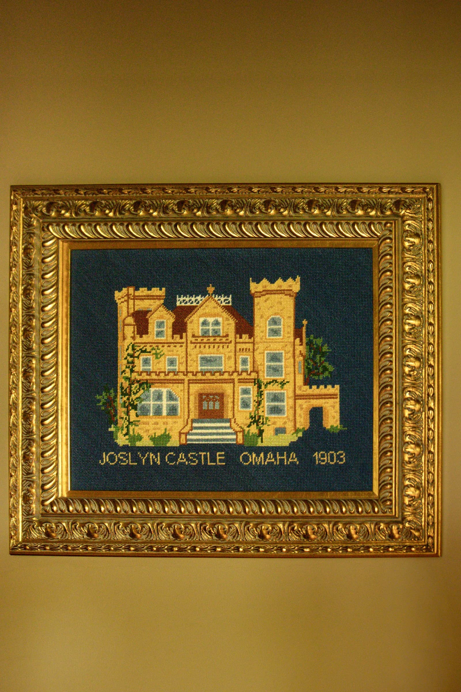 A needlepoint of the castle in a gilt frame, stitched with the words Joslyn Castle, Omaha, 1903.