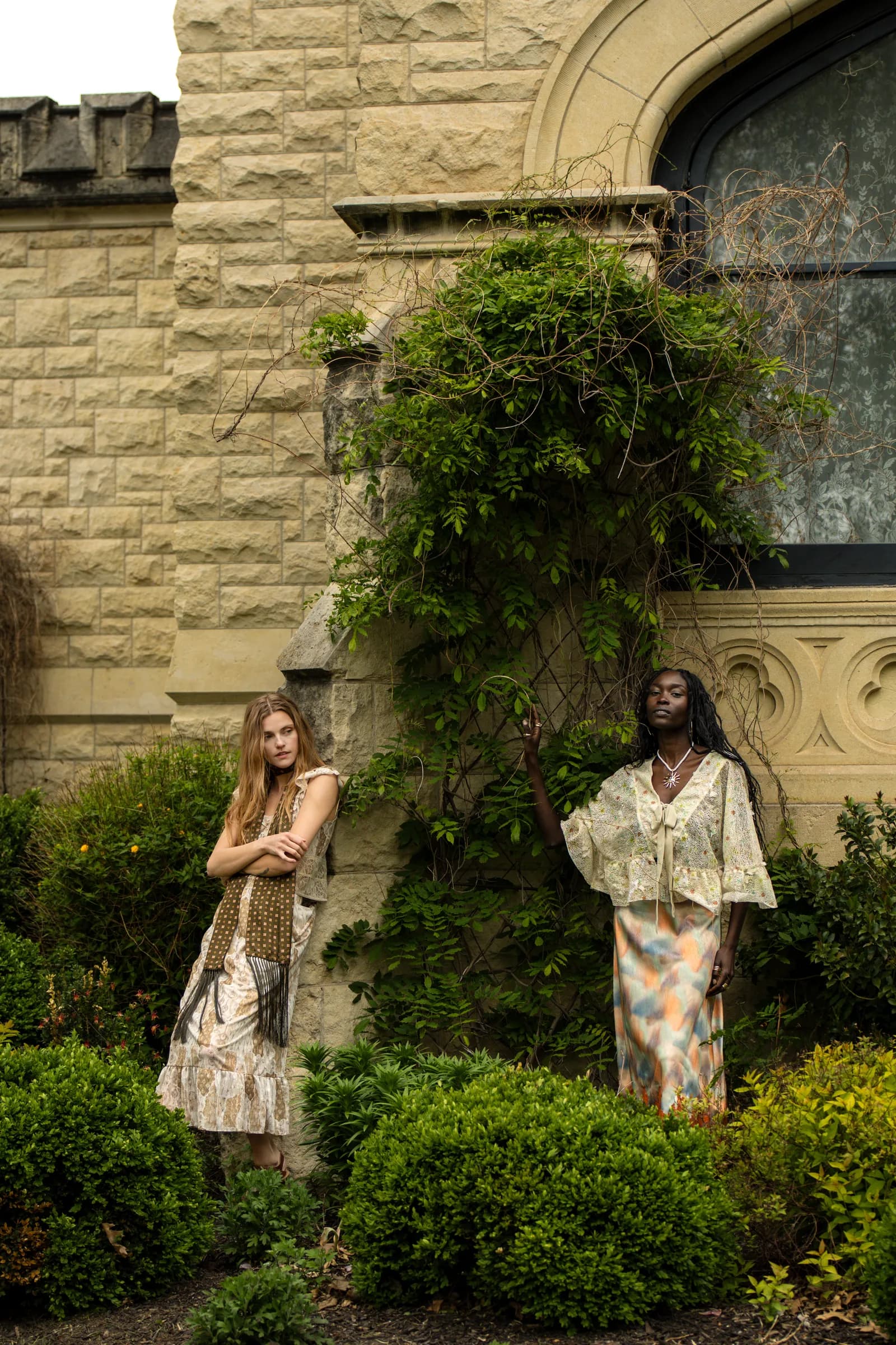 Two women stand apart in the climbing ivy and green of the castle garden.
