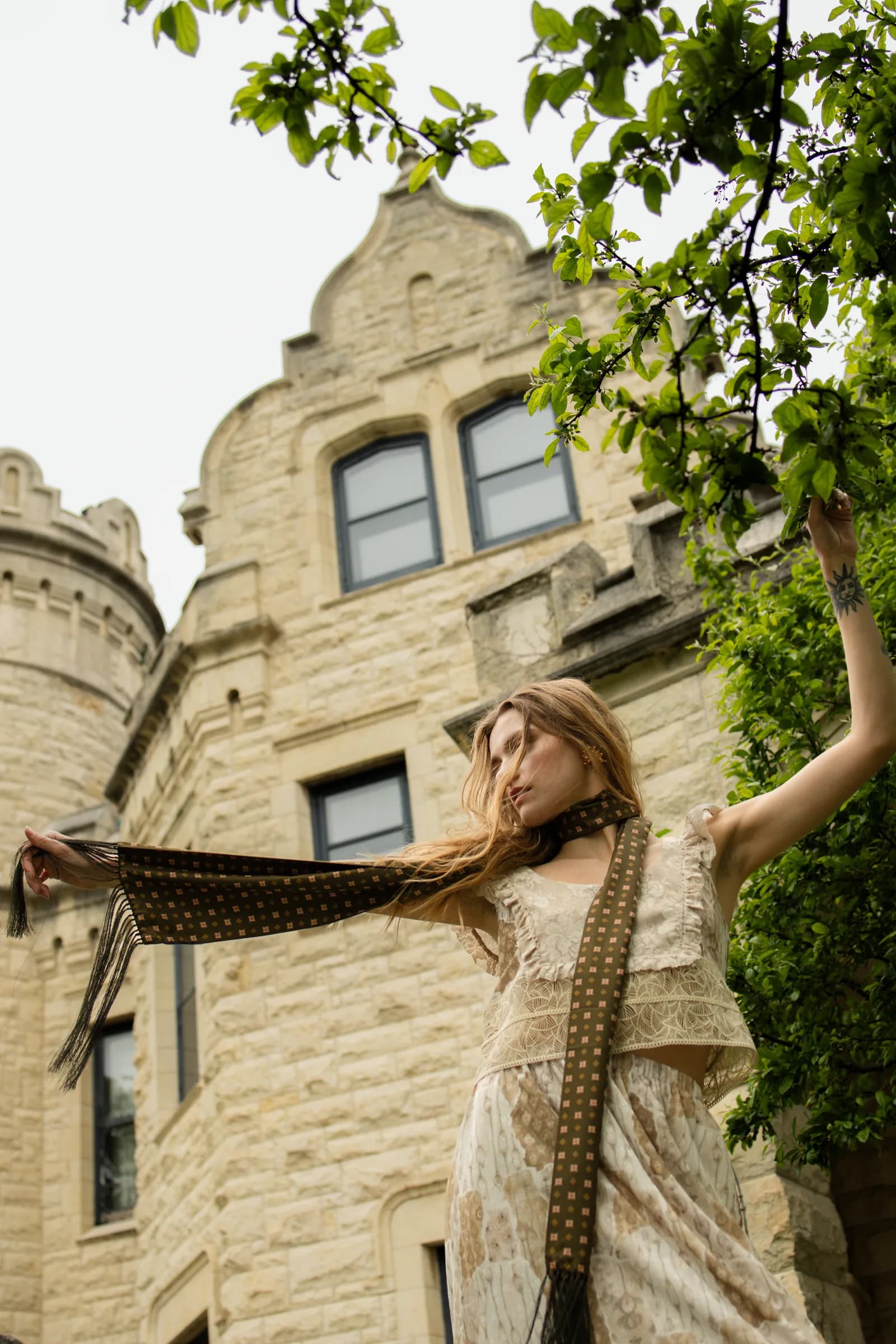 A woman lifts a long dotted scarf in the wind beneath a stone tower.
