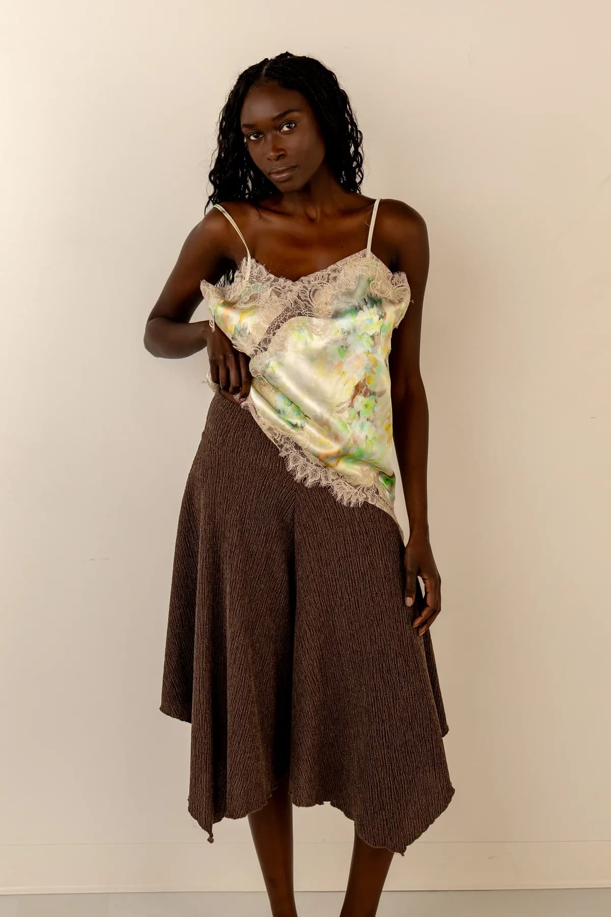 a woman on a cream studio backdrop in a watercolour satin cami with lace trim over a brown knit skirt with a pointed handkerchief hem