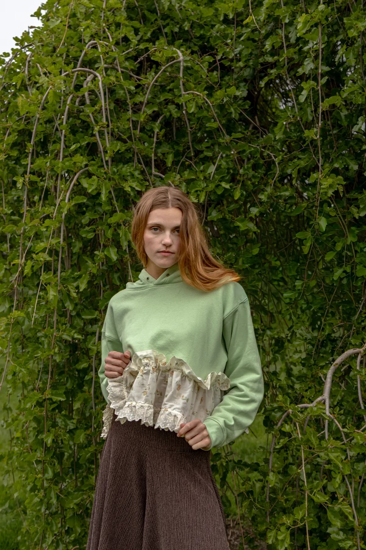 a woman in a green garden in a mint cropped hoodie with a lace ruffle hem and a long brown skirt