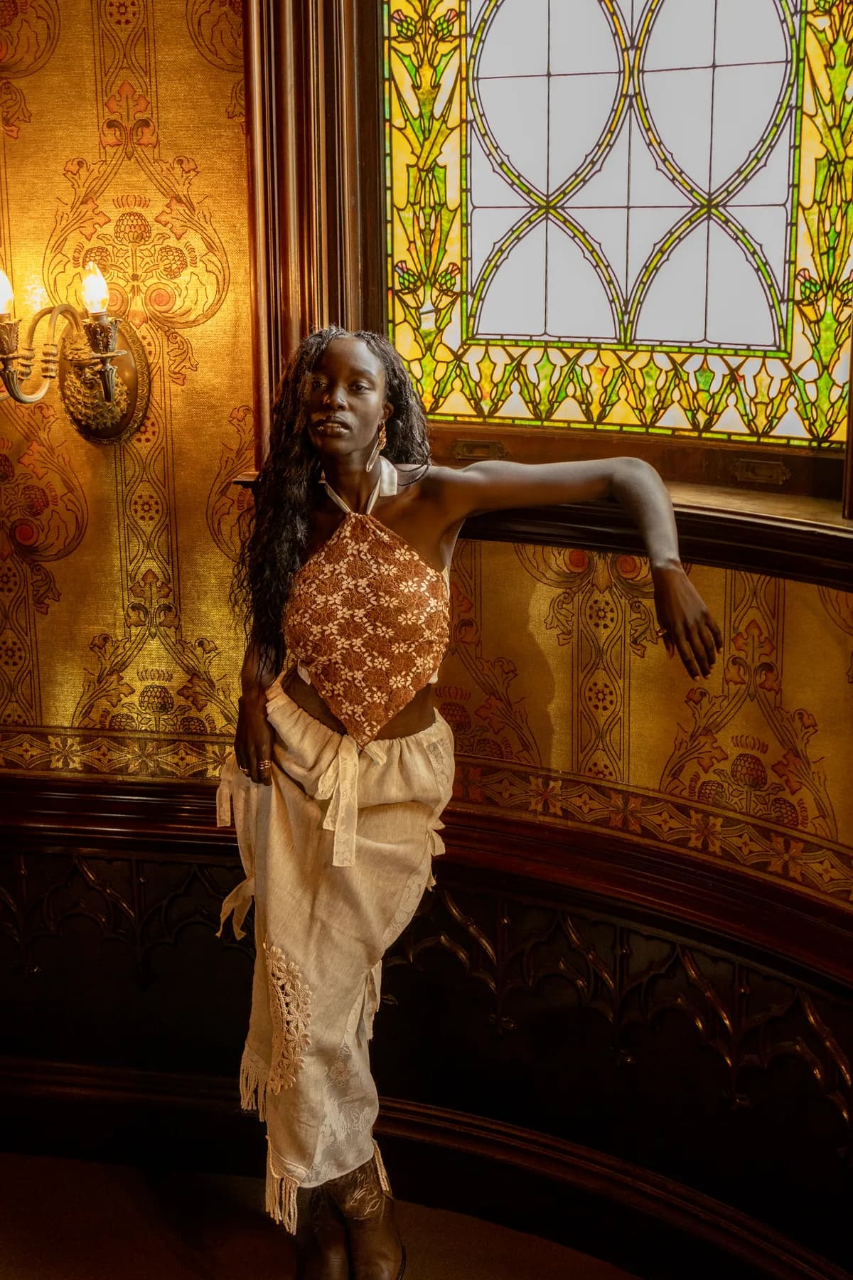 a woman under stained glass in a brown floral hanky halter and a cream statement skirt with crochet and fringe