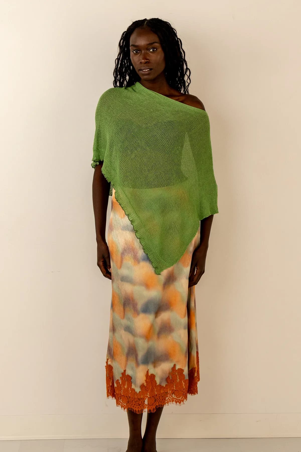 a woman on a cream studio backdrop in a sheer green knit poncho worn off one shoulder over a watercolour midi skirt with a rust lace hem