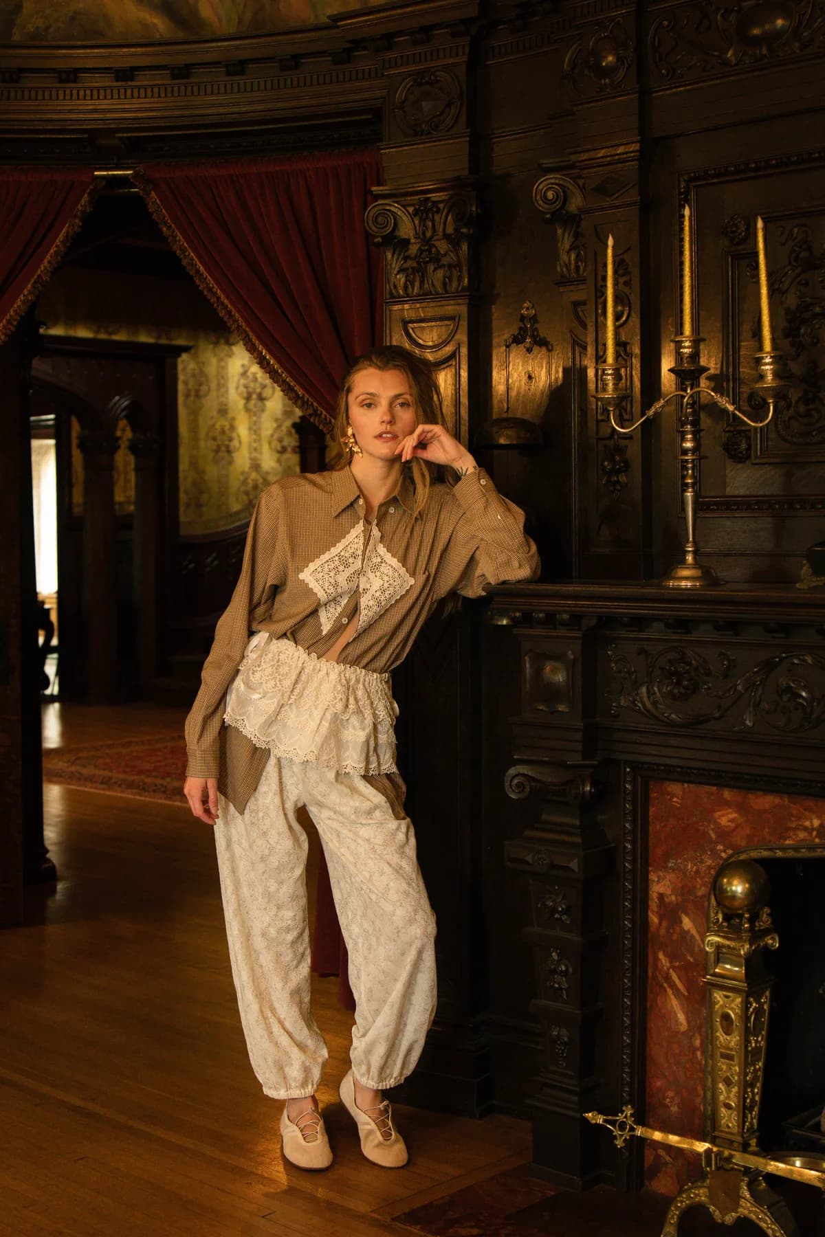 a woman by a carved fireplace in a brown button-front shirt with a lace patch over cream lace bubble pants